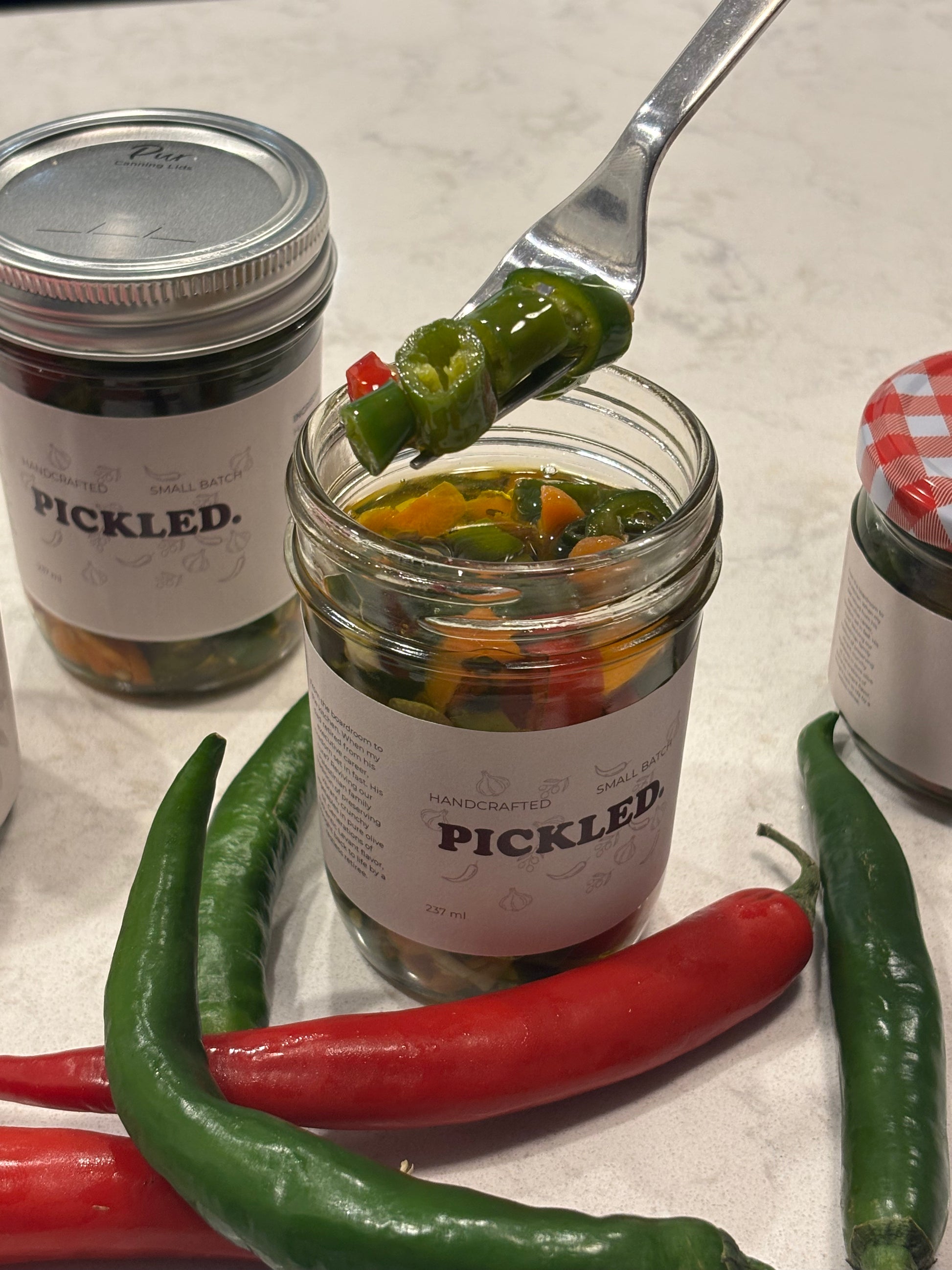 Jar of pickled vegetables with a spoonful being lifted, surrounded by fresh peppers on a countertop.